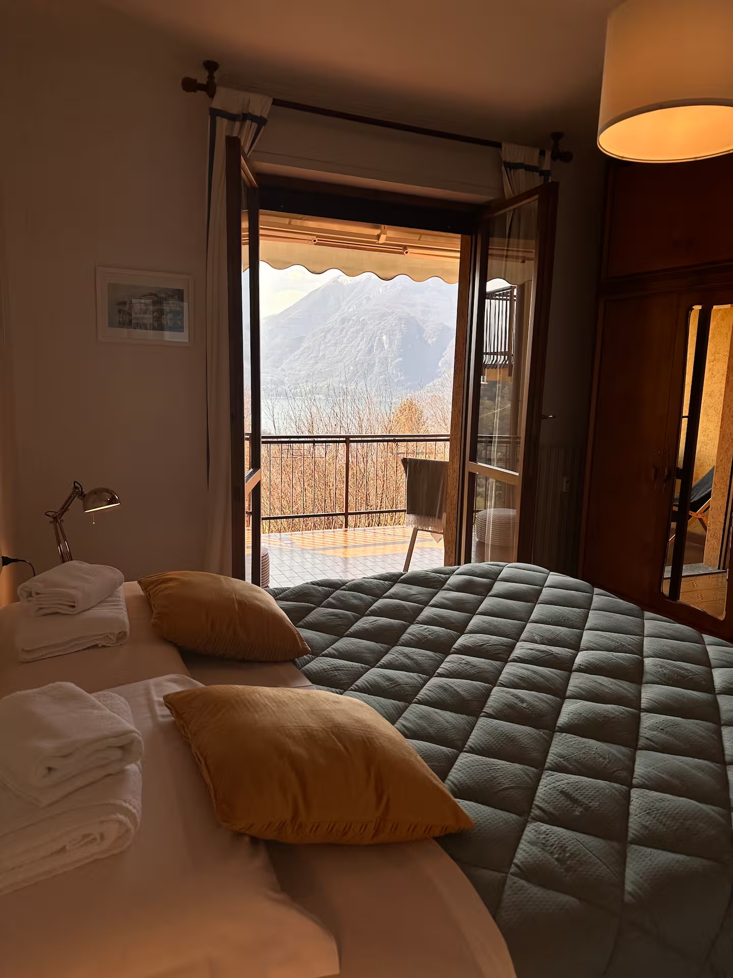 Double bedroom with a sage-green quilted bedspread, folded towels and mustard cushions, opening onto a balcony above the lake and mountains